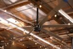 Cool Boss HVLS industrial ceiling fan installed in a metal-roof warehouse with visible steel trusses and electrical cabling