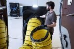 Technician connecting a yellow Cool Boss Ducting Kit (5150236) to a CB-16 evaporative cooler for aircraft pre-flight cooling inside a hangar. Another Cool Boss unit is visible in the background