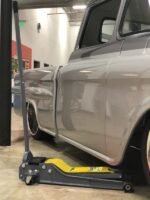 Low-profile gray hydraulic floor jack positioned under the side of a silver classic pickup truck inside a modern garage. The jack features a yellow saddle plate and long handle for easy lifting